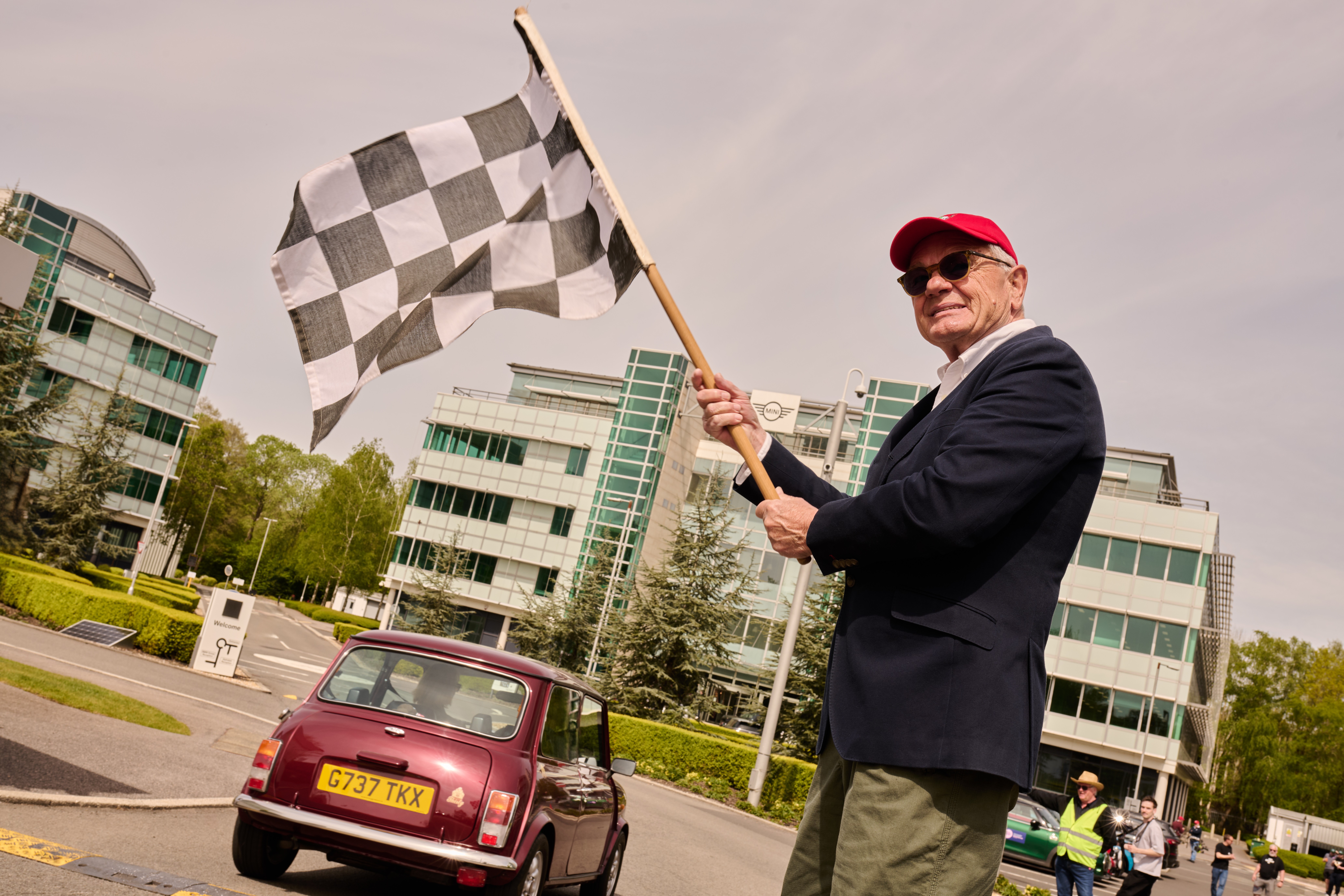 A silver Mini Cooper, adorned with Union Jack mirrors, speeds down a scenic road, followed closely by red and blue Minis among green blurred trees.